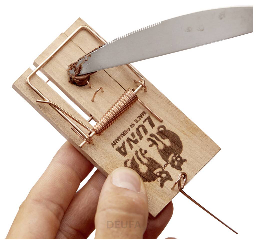 Eine Hand hält eine Mausefalle, während ein Messer ihren Auslöser auslöst und eine gefährliche Anordnung auf einer Holzfalle mit der Aufschrift 