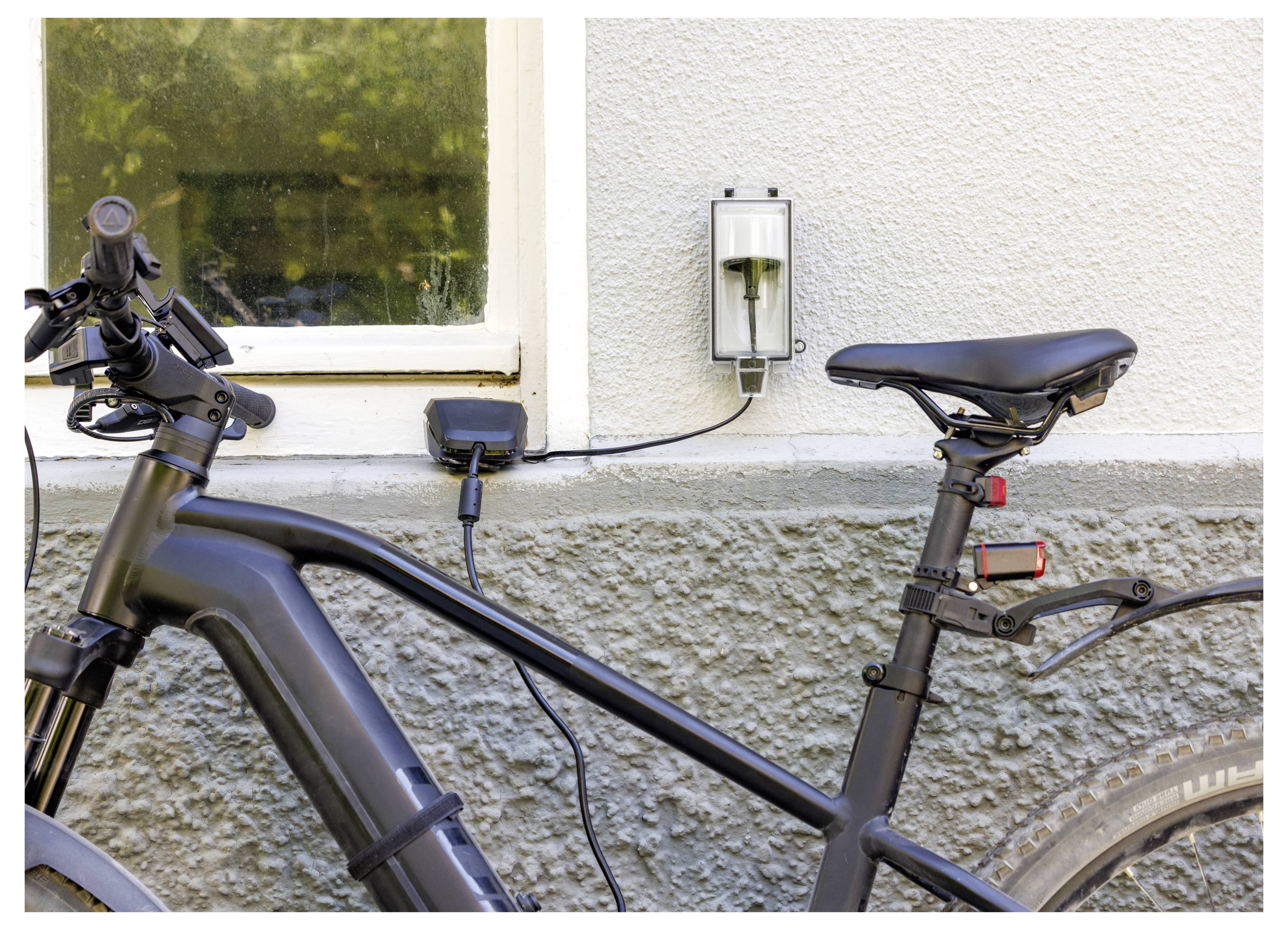 Elektrisches Fahrrad, das an einer wandgestützten Ladestation neben einem Fenster angeschlossen ist und zeigt, dass das Fahrrad im Freien aufgeladen wird.