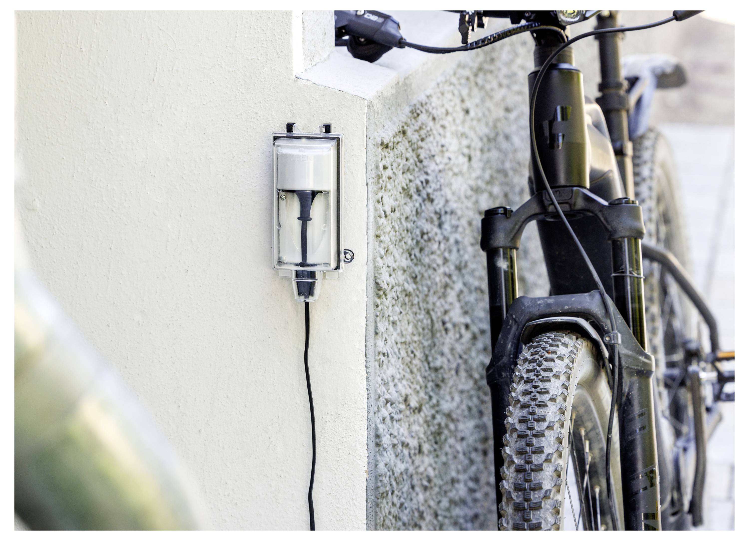 Wandmontiertes Elektrofahrrad-Ladegerät neben einem geparkten Mountainbike, mit einem Kabel, das mit dem Fahrradrahmen verbunden ist.