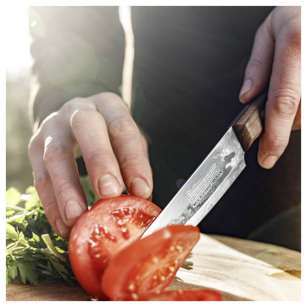 Hände schneiden eine Tomate auf einem Schneidebrett mit einem Messer, mit Kräutern daneben und warmem Sonnenlicht im Hintergrund.