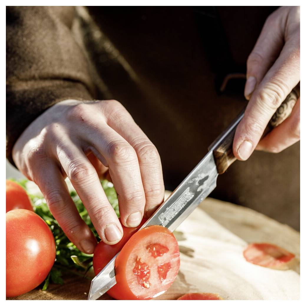 Hände schneiden eine Tomate mit einem Messer auf einem Schneidebrett, während frisches Gemüse zubereitet wird.