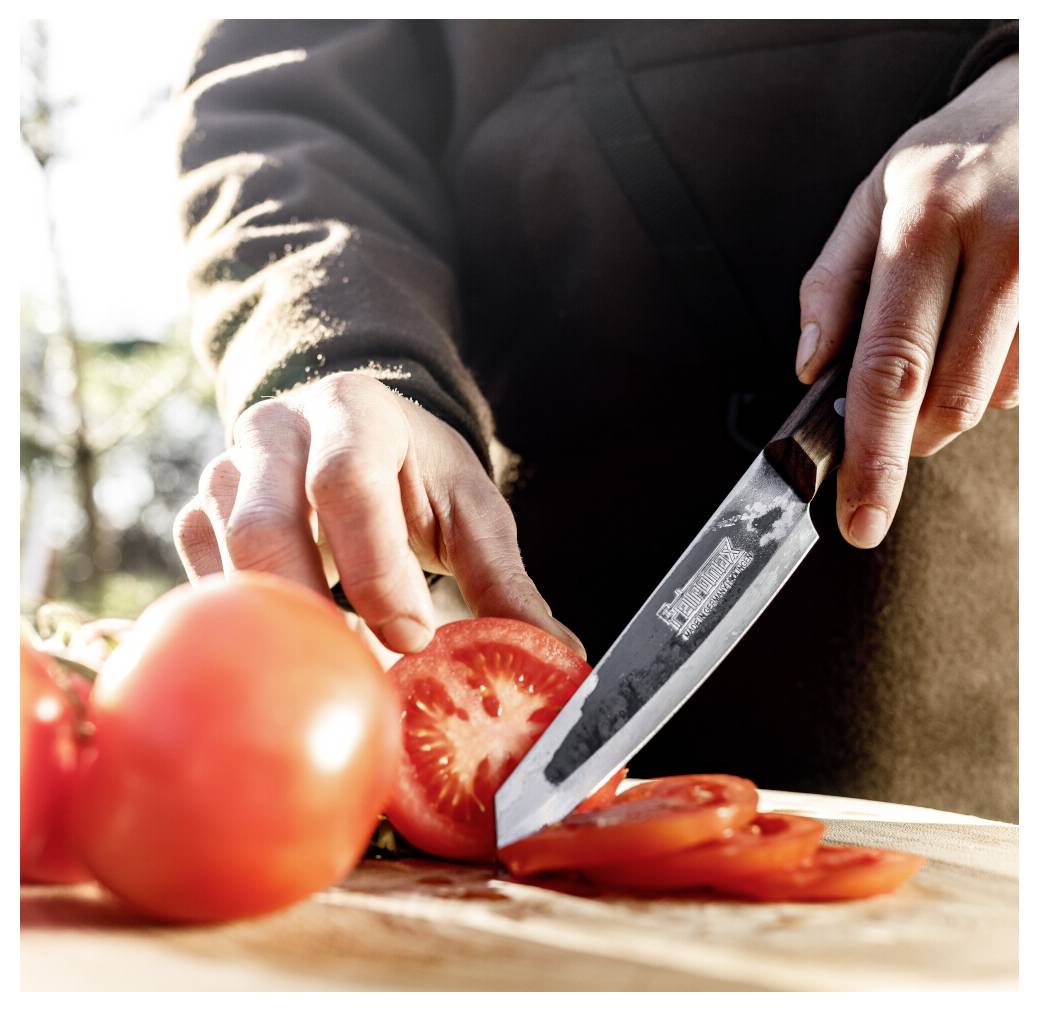 Eine Person schneidet eine Tomate mit einem Kochmesser auf einem Schneidebrett im Freien bei hellem Sonnenlicht.