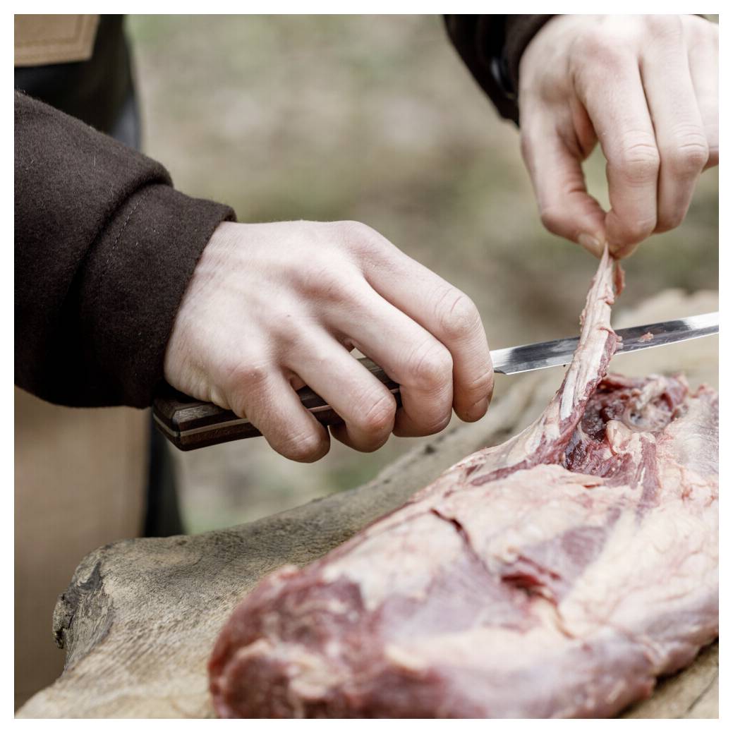 Hände, die ein Messer verwenden, um Fleisch auf einer Schneidfläche im Freien zu trimmen.