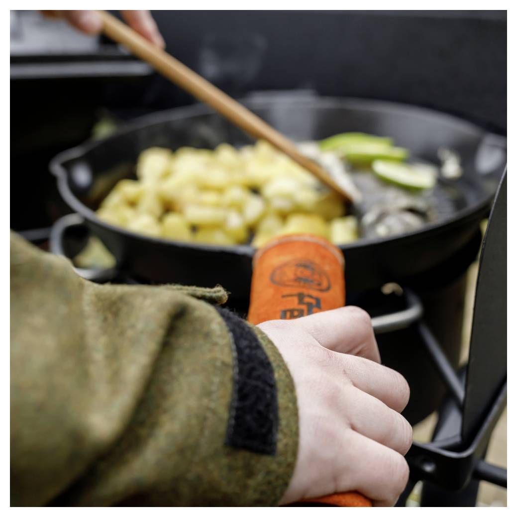 Eine Person rührt Essen in einer Pfanne über einem Campingkocher um und hält dabei ein Küchengerät mit orangefarbenem Griff im Freien.