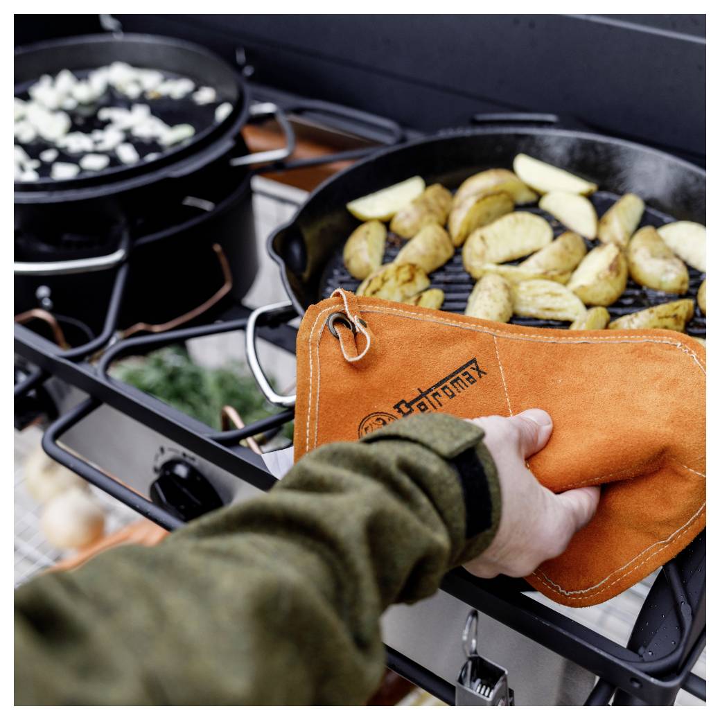 Hand mit einem orangefarbenen Ofenhandschuh, der eine Pfanne mit gebratenen Kartoffelecken auf einem Außengrill bewegt; ein Topf siedelt daneben.
