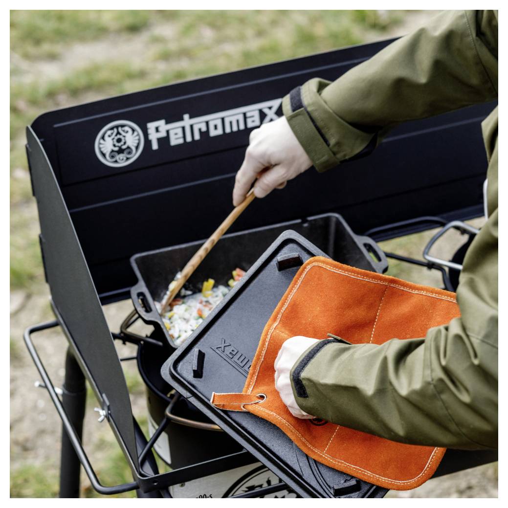 Person bedient einen Petromax-Campingkocher und verwendet einen Pfannenwender sowie einen orangefarbenen Ofenhandschuh, um eine heiße Gusseisenpfanne mit Essen im Freien zu handhaben.