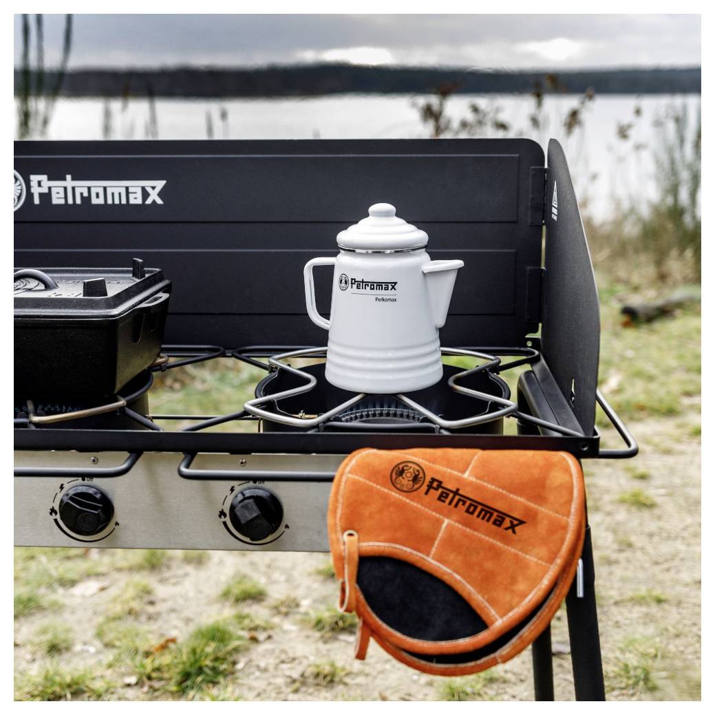 Campingkocherstützung mit einem weißen Petromax-Kessel auf dem Brenner und einem orangefarbenen Petromax-Ofenhandschuh, der vorne hängt, im Freien an einem See.