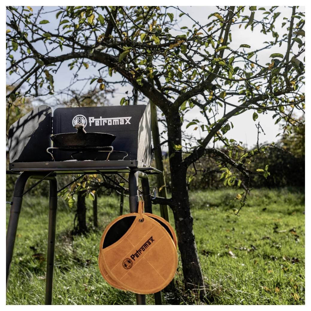 Tragbarer Petromax-Campingkocher auf einem Ständer im Freien unter einem Baum, mit einer Pfanne oben drauf und einer orangefarbenen Petromax-Tasche, die darunter hängt.