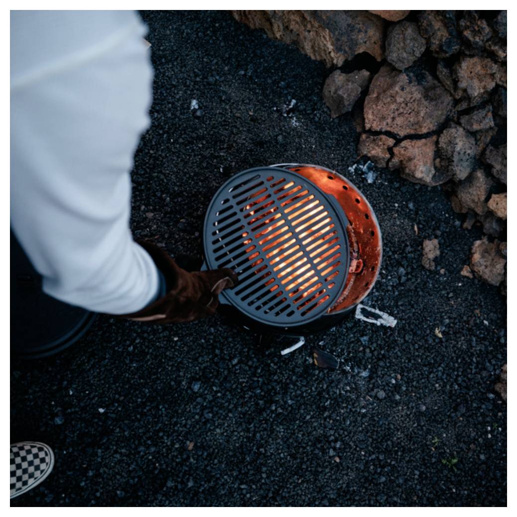 Person, die einen kleinen glühenden Holzkohlegrill im Freien auf dunklem Kies in der Nähe von Steinen bedient.