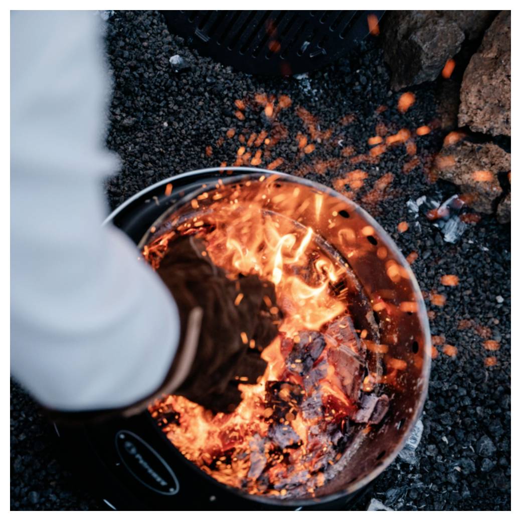 Person, die ein Lagerfeuer oder eine Feuerstelle betreut, wobei glühende Glut in einem Metallbehälter mit sprühenden Funken auf Kies im Freien umgerührt wird.