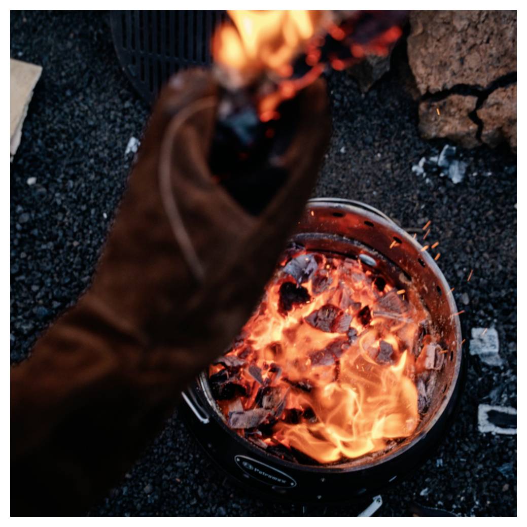Eine behandschuhte Hand streichelt ein helles Lagerfeuer in einer Metallfeuergrube auf dunklem Kies.