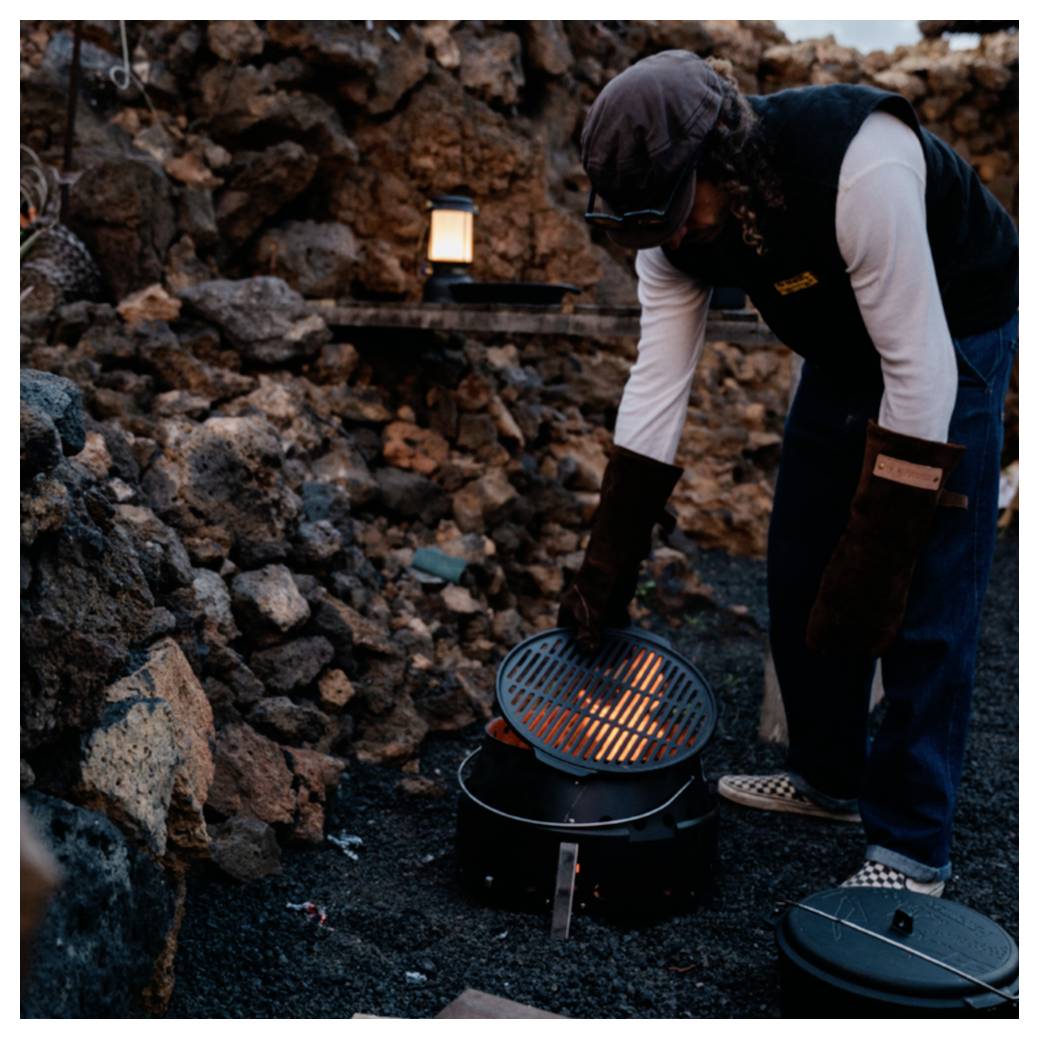 Person, die einen kleinen Holzkohlengrill im Freien zwischen Steinen bedient, trägt Handschuhe und lehnt sich über den Grill in der Nähe eines von einer Laterne beleuchteten Campingplatzes.