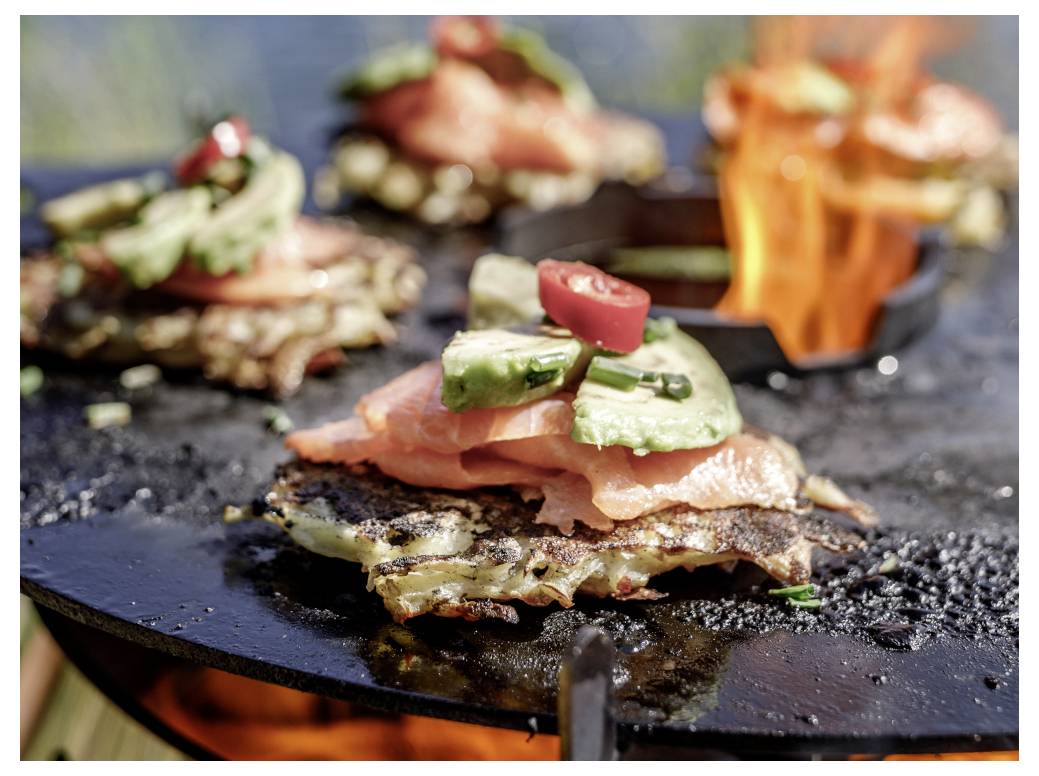 Lachs- und Avocado-belegte Frikadellen zischen auf einer runden Grillplatte über einer offenen Flamme an einem Außengrill.