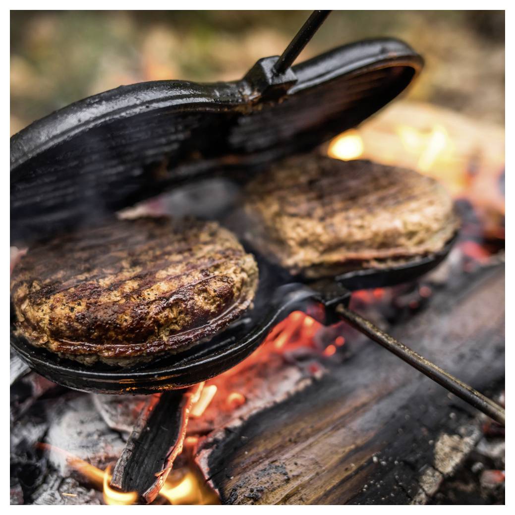 Zwei Burger braten in einer gusseisernen Pfanne über glühenden Lagerfeuerkohlen, wobei der Deckel teilweise offen ist.