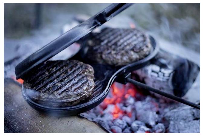 Zwei Hamburger-Pattys grillen in einer gusseisernen Pfanne über glühenden Kohlen