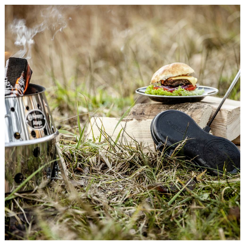 Outdoor-Kochausrüstung mit einem Holzkohlengrill, einem Burger auf einem Teller und einer Gusseisenpfanne auf einer grünen Wiese.
