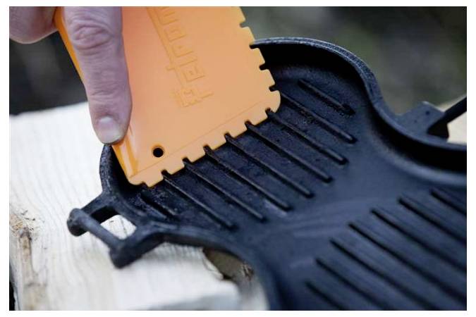 Eine Hand benutzt einen orangen Schaber, um eine schwarze gusseiserne Plancha oder Pfanne auf einer Holzoberfläche zu reinigen.