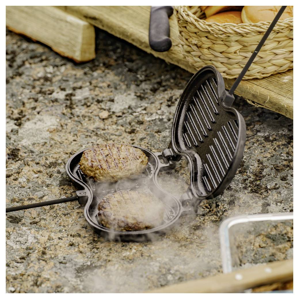 Zwei Burger werden in einer kleeblattförmigen Grillpfanne mit offnem Deckel gekocht, Dampf steigt auf einer Steinoberfläche im Freien auf.
