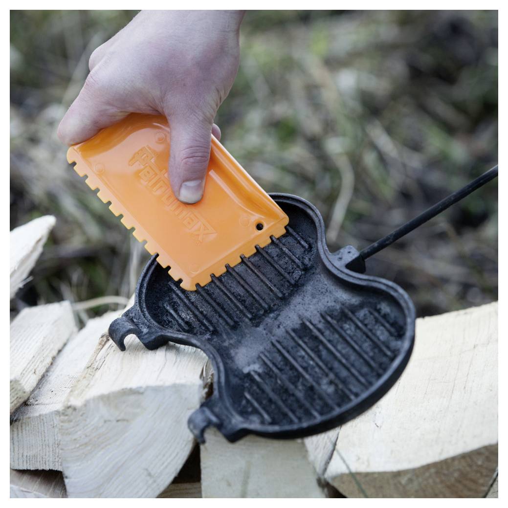 Hand mit einem orangefarbenen Feueranzünder, um einen schwarzen Metall-Feuer­schläger auf gespaltenes Holz im Freien zu entzünden.