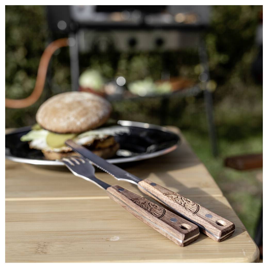 Gegrillter Burger auf einem schwarzen Teller im Freien, mit Gabel und Messer auf einem Holztisch im Vordergrund.
