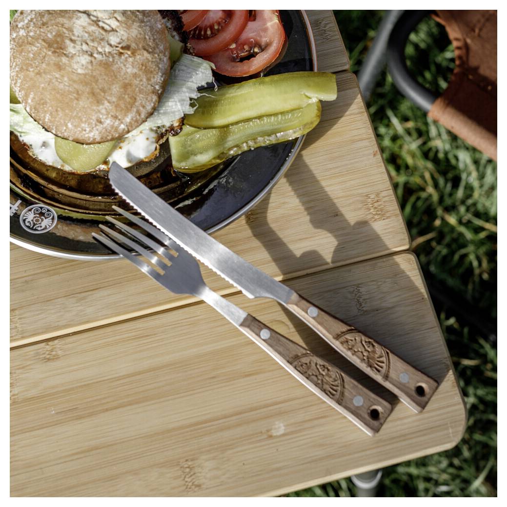 Ein halb aufgegessener Burger auf einem schwarzen Teller mit Gurken und Tomate, ruht auf einem Holzschneidebrett neben einer Gabel und einem Messer im Freien.