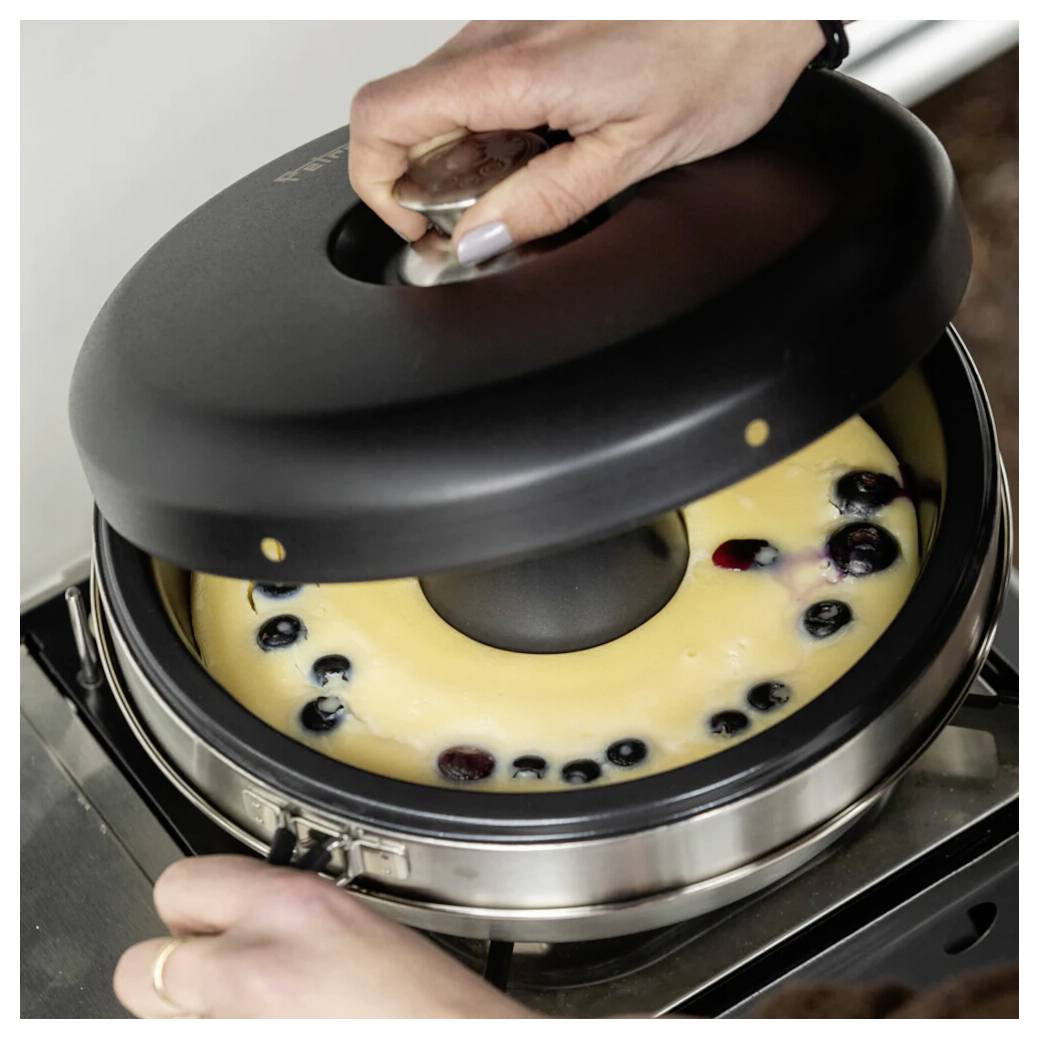 Eine Hand hebt den Deckel einer Käsekuchen-Springform an und offenbart eine helle Füllung, die mit Blaubeeren belegt ist und einen dunklen Mitteleinsatz hat.