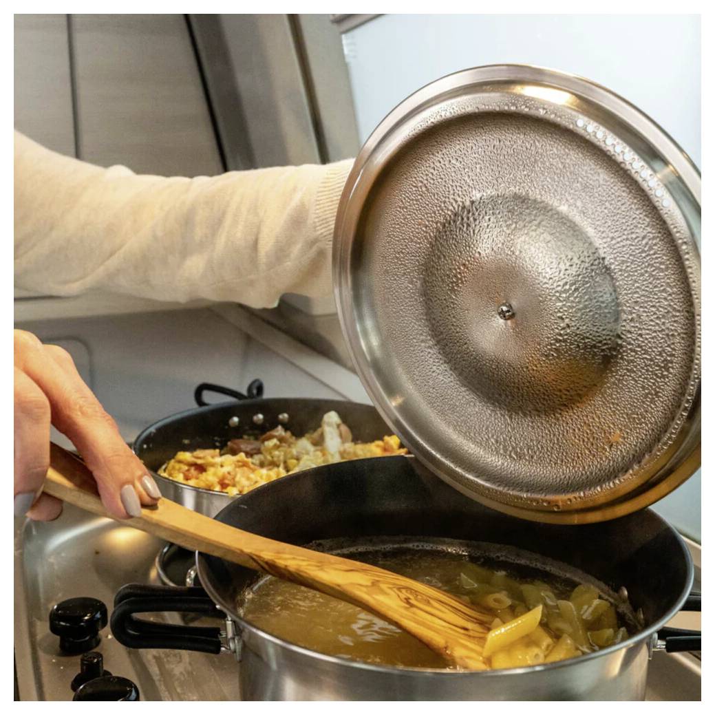 Eine Person hebt einen Topfdeckel an, während sie Suppe auf einem Herd mit einem Holzlöffel umrührt; eine weitere Pfanne mit Essen kocht in der Nähe.