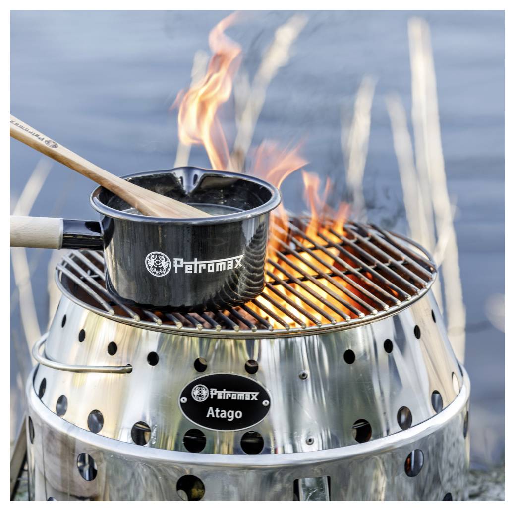 Kochtopf zum Camping mit Holzlöffel sitzt über einer offenen Flamme auf einem Grill aus Edelstahl von Petromax Atago am Wasser, mit sichtbarem Petromax Atago-Branding.