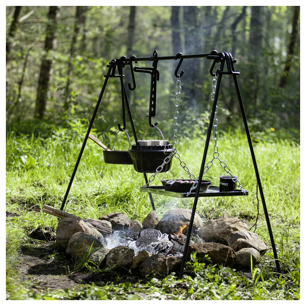 Outdoor-Lagerfeuer-Kochaufbau mit einem Dreibein, hängenden Töpfen und einem Grill über Steinen und Brennholz in einer Waldlichtung.