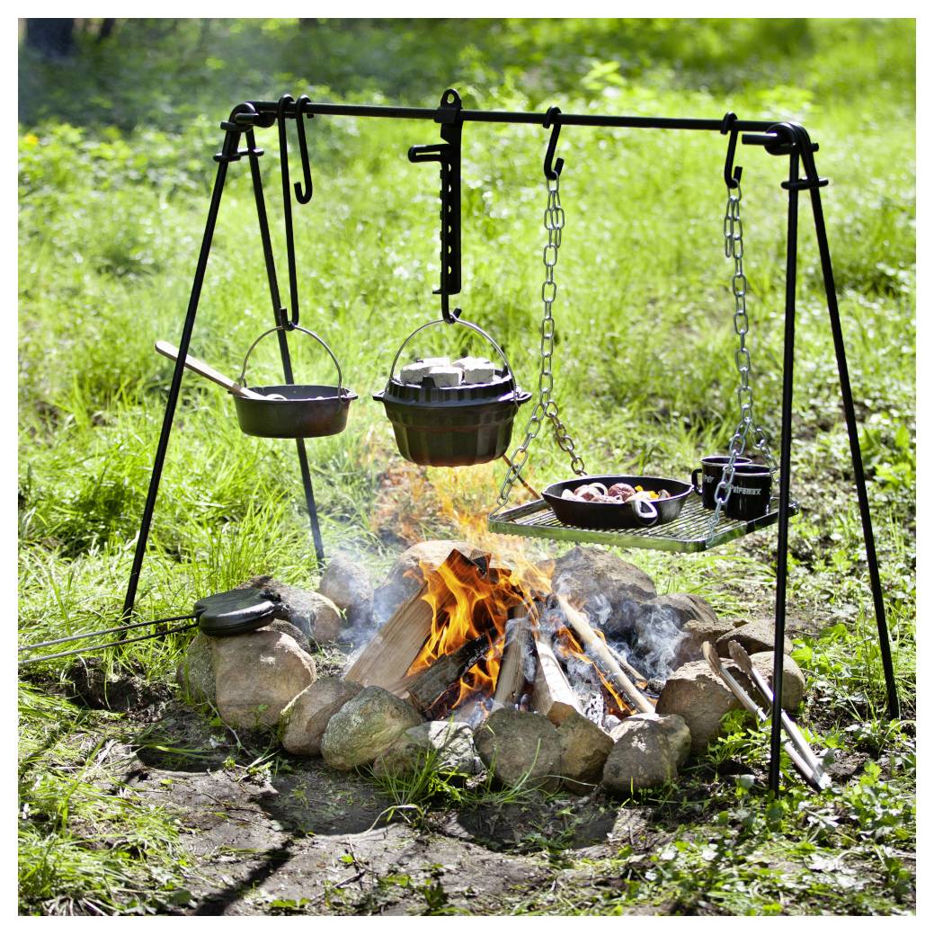 Outdoor-Lagerfeuer-Kochaufbau mit einem Dreibein und hängenden Töpfen über einem Feuer, umgeben von Steinen und Gras.