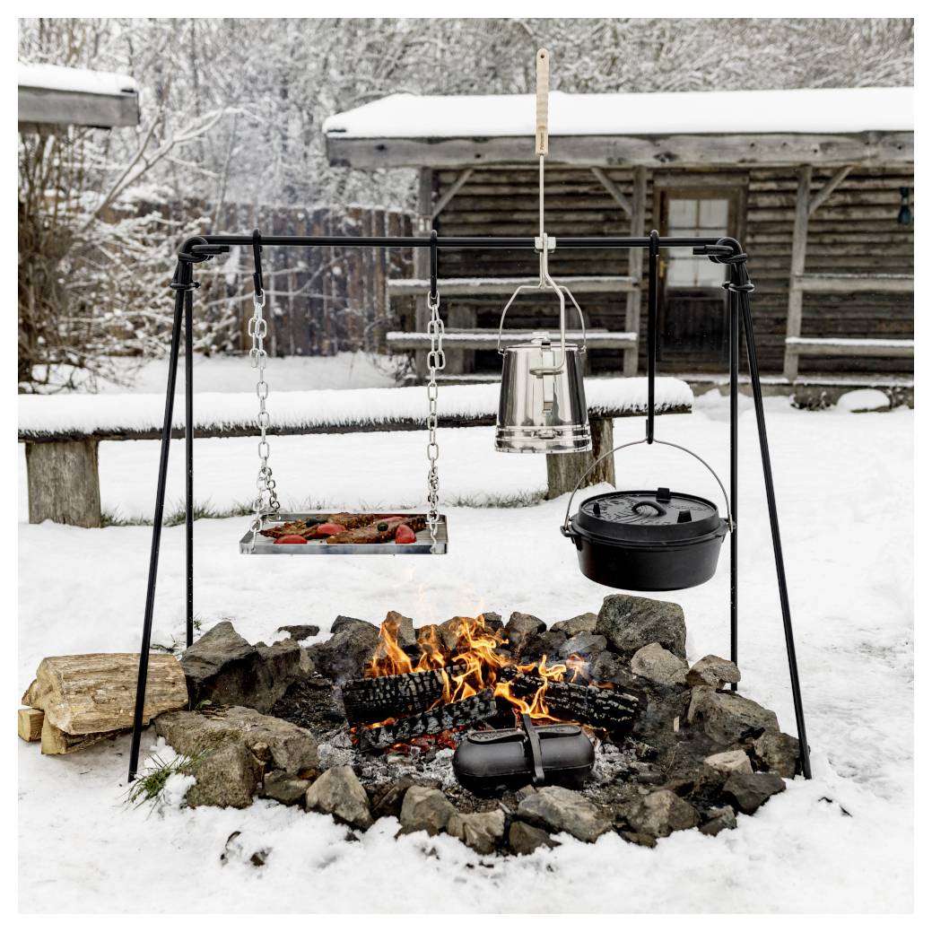 Schneebedeckter Outdoor-Lagerfeuer-Kochplatz mit hängender Grillschale, Kessel und gusseisernem Topf über einem offenen Feuer neben einer Blockhütte.