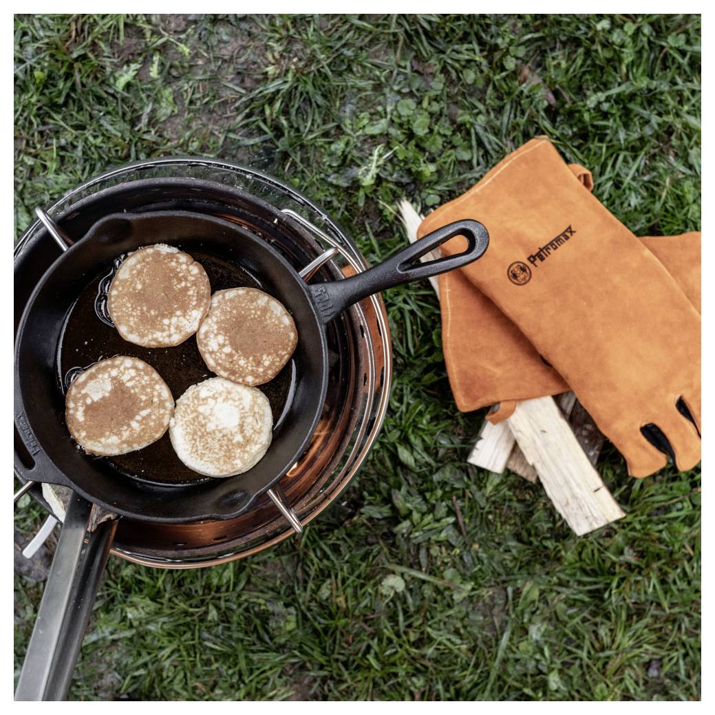 Pfannkuchen, die in einer Gusseisenpfanne auf einem Campingkocher auf Gras kochen, mit orangefarbenen Ofenhandschuhen und Brennholz in der Nähe.