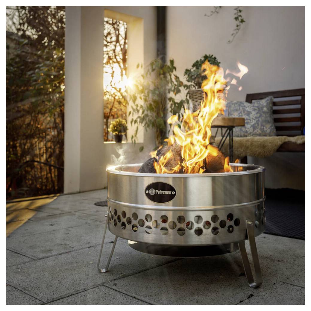 Ein Feuertisch aus Edelstahl mit hohen Flammen, die auf einer Terrasse neben Outdoor-Möbeln bei Sonnenuntergang brennen.