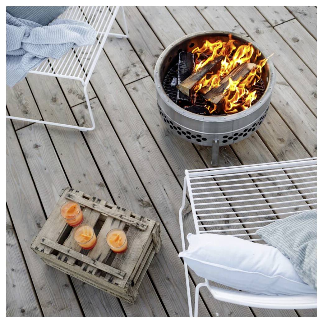 Terrassendeck mit einer beleuchteten Feuerstelle, zwei weißen Liegestühlen mit Kissen und Decken sowie einem Holztablett mit drei orangefarbenen Kerzen.