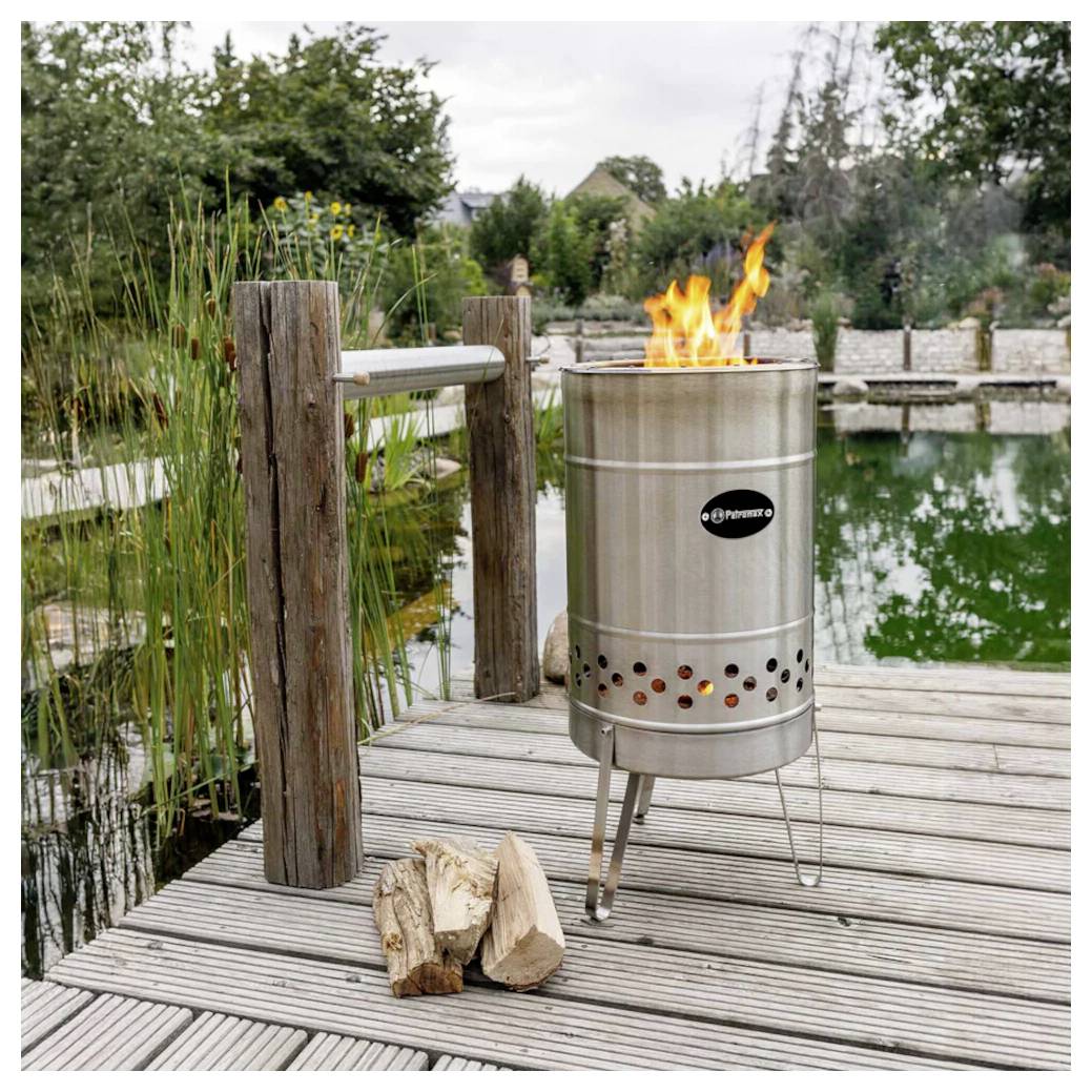 Rostfreier Stahl-Außenfeuerstelle, die auf einem hölzernen Steg neben einem Teich brennt, mit gehacktem Brennholz in der Nähe und einem Garten im Hintergrund.