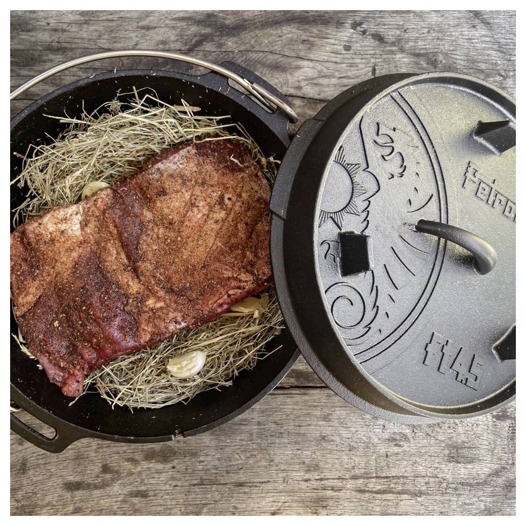 Geräuchertes Fleischbrisket in einem gusseisernen Bräter, mit Stroh ausgelegt, Deckel offen, auf einer Holzoberfläche.
