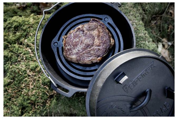Steak wird in einem gusseisernen Dutch Oven im Freien gekocht, wobei der Deckel neben dem Topf auf dem Gras liegt.