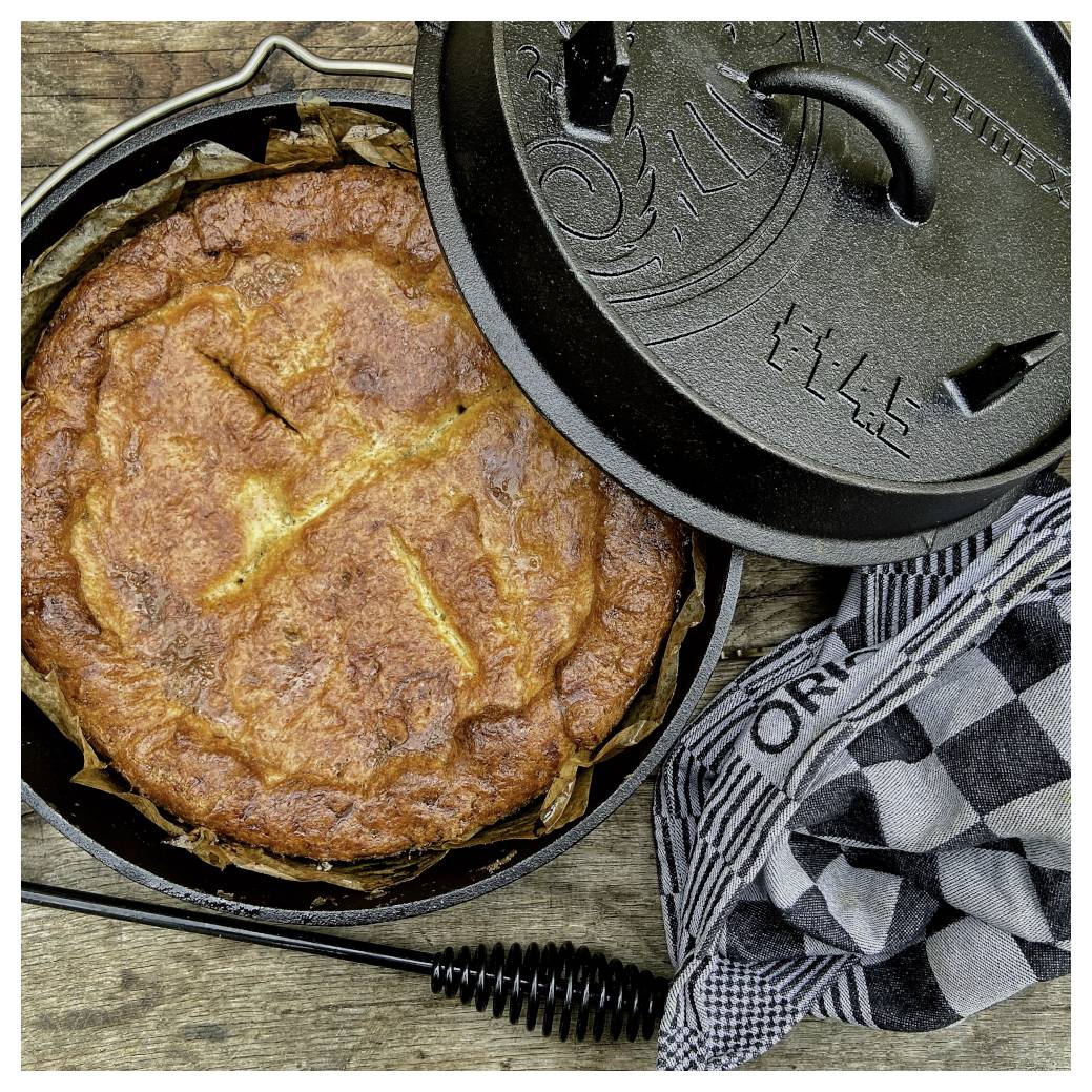 Frisch gebackenes rundes Brot oder eine Pie in einem gusseisernen Dutch Oven mit offenenem Deckel auf einer Holzoberfläche, neben einem karierten Küchentuch.