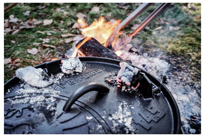 Ein gusseiserner Dutch-Oven-Deckel sitzt neben einem Lagerfeuer, mit glühenden Kohlen und einem Metallwerkzeug, das heiße Glut im Freien hebt.