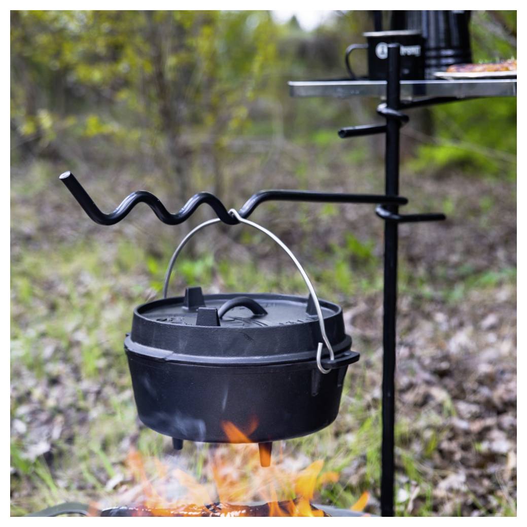 Schwarzer gusseiserner Topf hängt über einem Lagerfeuer auf einem dreibeinigen Grillgestell im Freien, mit sichtbaren Flammen darunter.