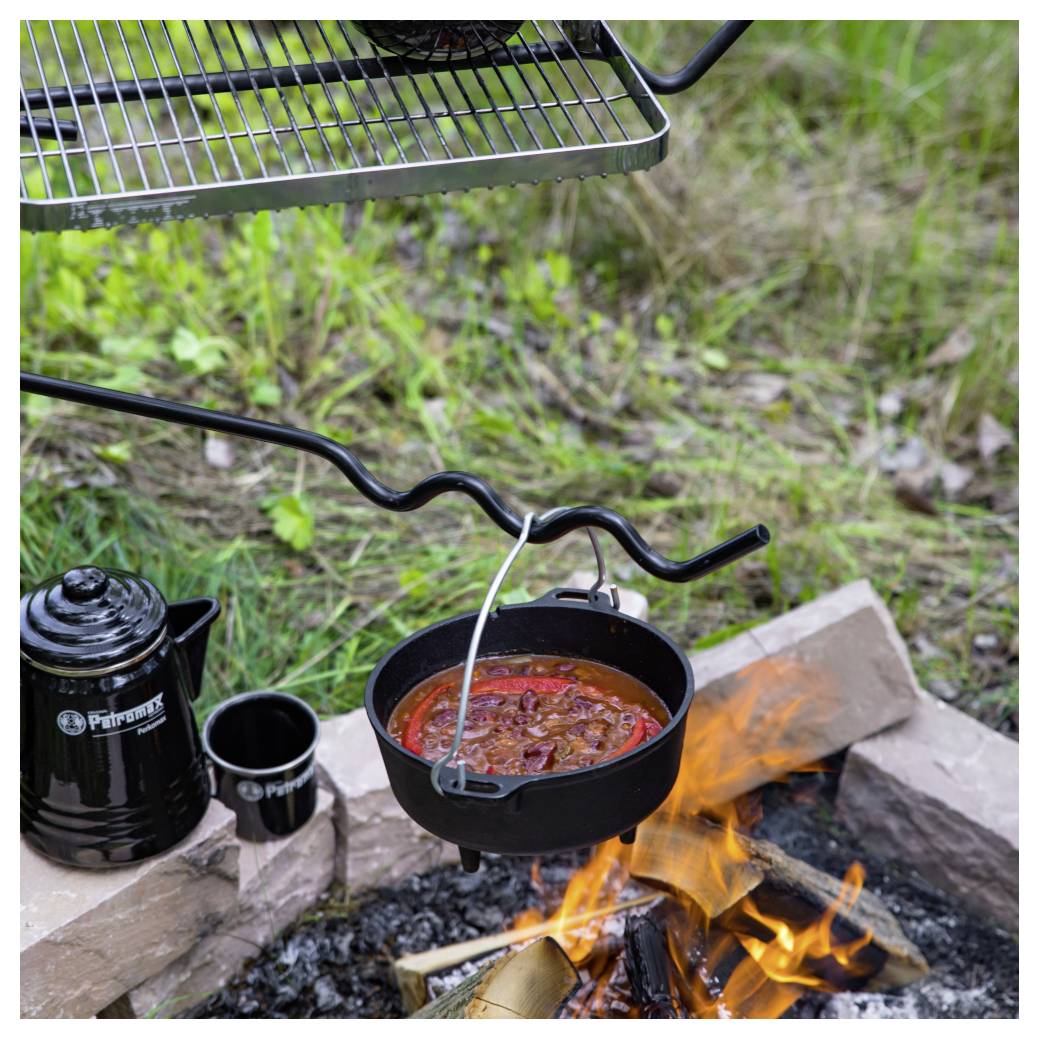 Ein gusseiserner Topf mit rotem Eintopf hängt über einem Lagerfeuer neben einer Kaffeekanne und einer Tasse, mit einem Grillrost darüber.