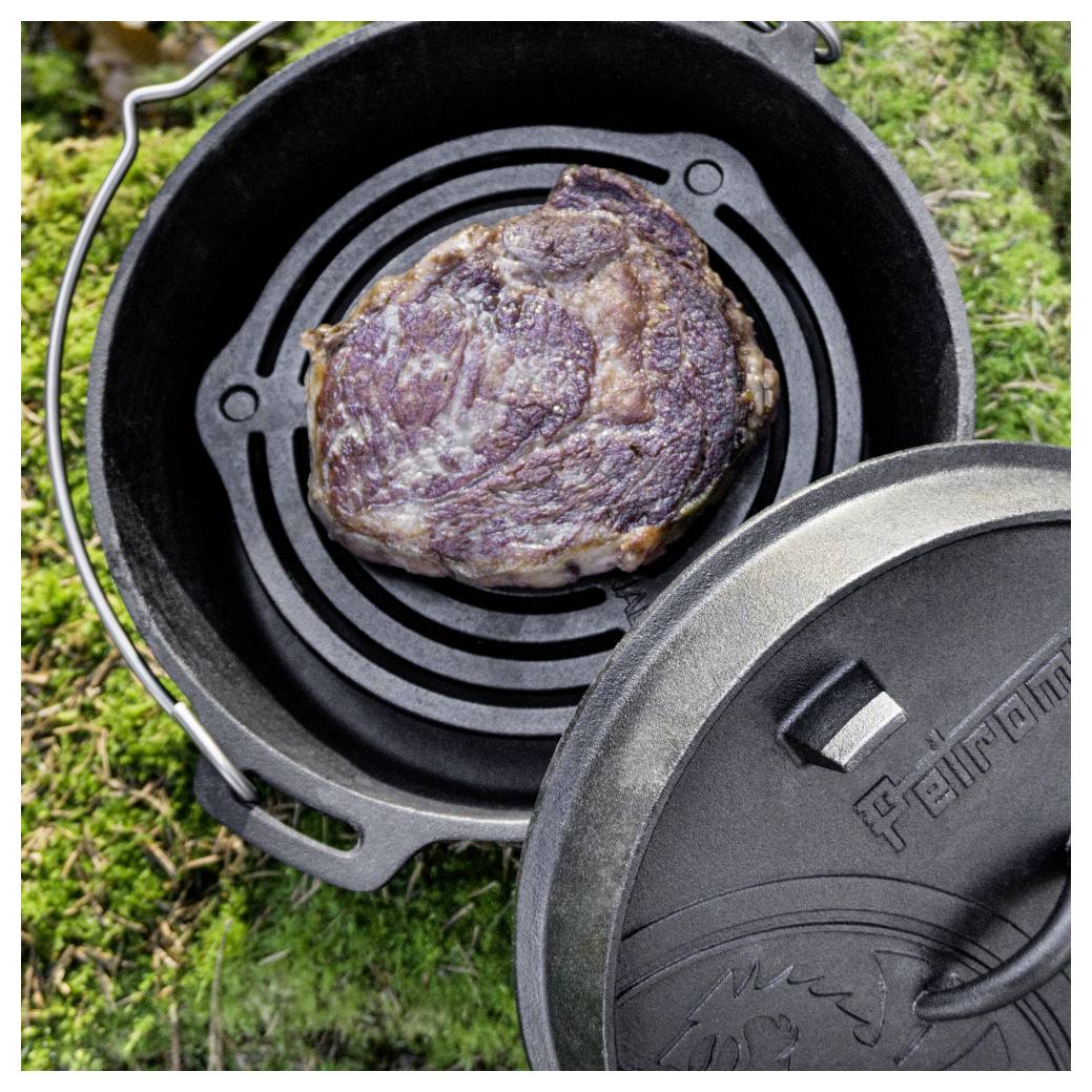 Ein gegrilltes Steak sitzt in einem gusseisernen Schmortopf im Freien auf Gras, wobei der Deckel teilweise offen ist.