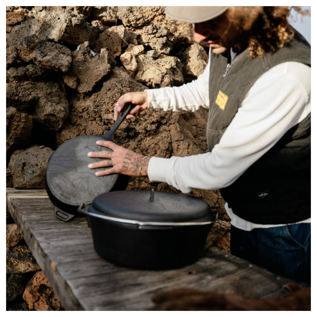 Person im Freien öffnet einen schwarzen Dutch Oven auf einem Holztisch neben einer felsigen Steinmauer.