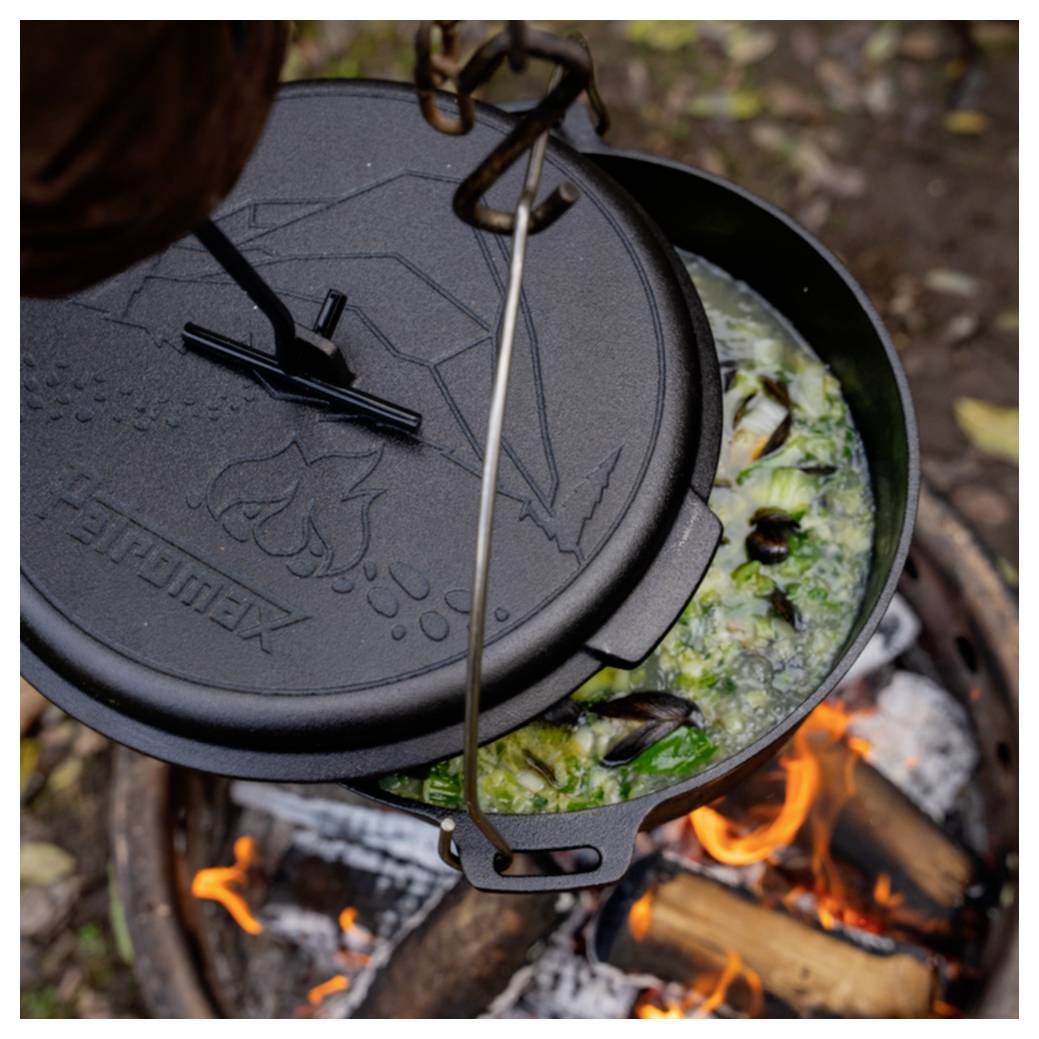 Dutchoven mit angehobenem Deckel über Lagerfeuer, das einen köchelnden Muschel- und Gemüseeintopf im Freien zeigt.