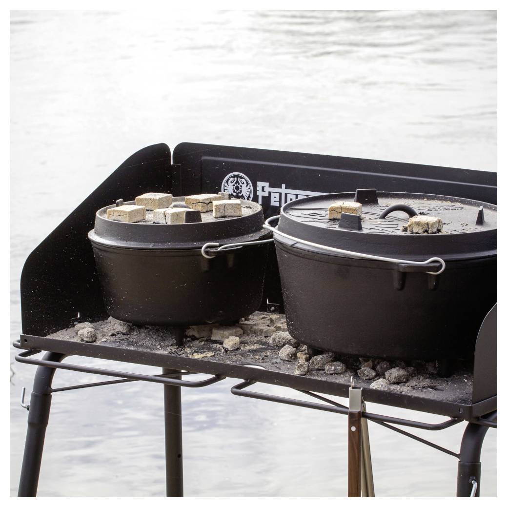 Zwei gusseiserne Töpfe, die auf einem tragbaren Outdoor-Kocher am Wasser köcheln, mit Glut und Holzstücken oben drauf.