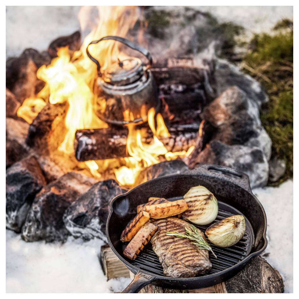 Steak, Zwiebeln und Süßkartoffeln, die in einer gusseisernen Pfanne über einem Lagerfeuer garen, mit einem Kessel, der dahinter in den Flammen erhitzt wird.