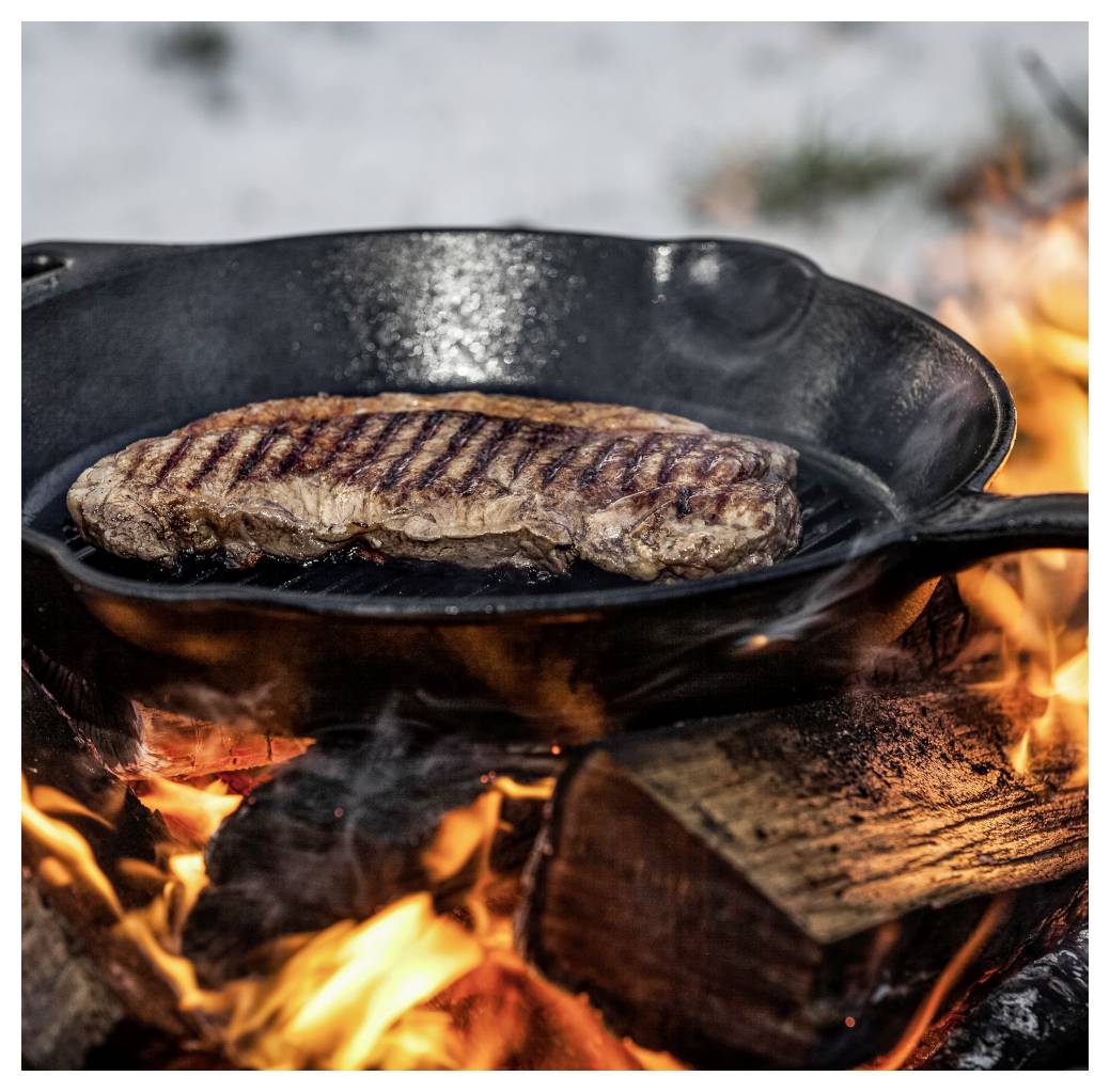 Steak wird in einer gusseisernen Pfanne über einem offenen Holzfeuer angebraten, mit Flammen und Rauch unter der Pfanne.