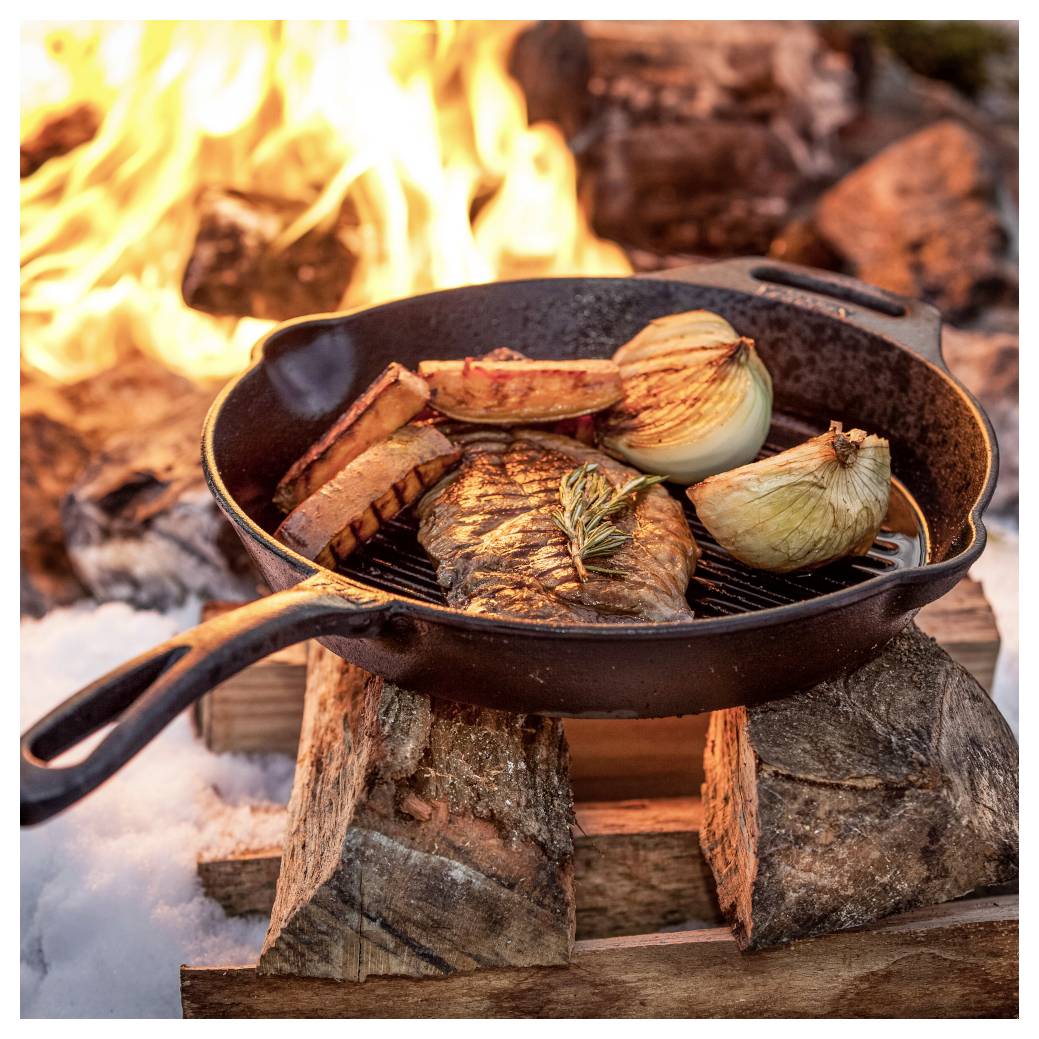 Gusseiserne Pfanne mit Steak, Kartoffeln und Zwiebeln, die über einem offenen Lagerfeuer auf gestapelten Holzscheiten und Schnee kochen.