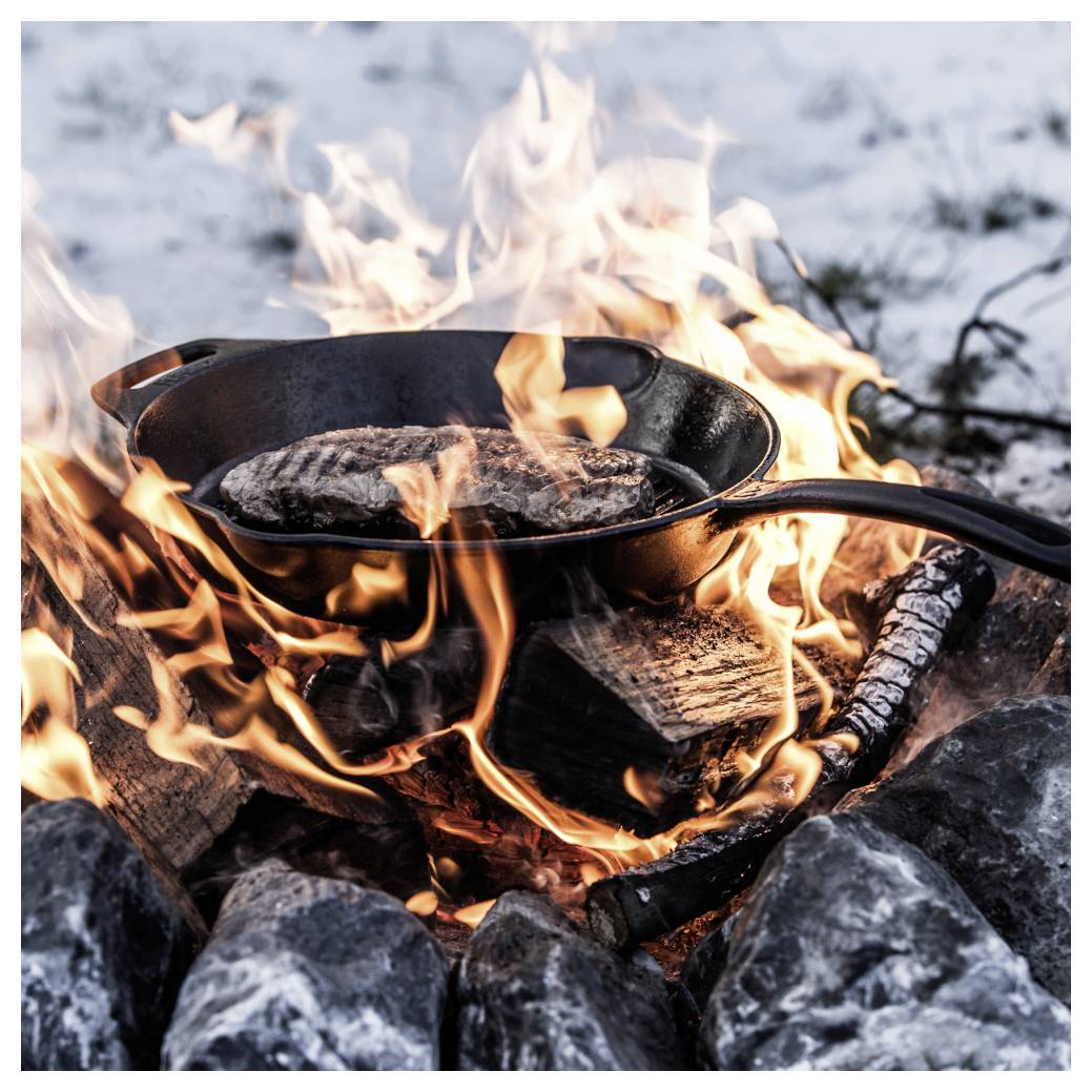 Eine gusseiserne Pfanne mit einem Steak, das über einem offenen Lagerfeuer kocht, umgeben von Steinen und Schnee.