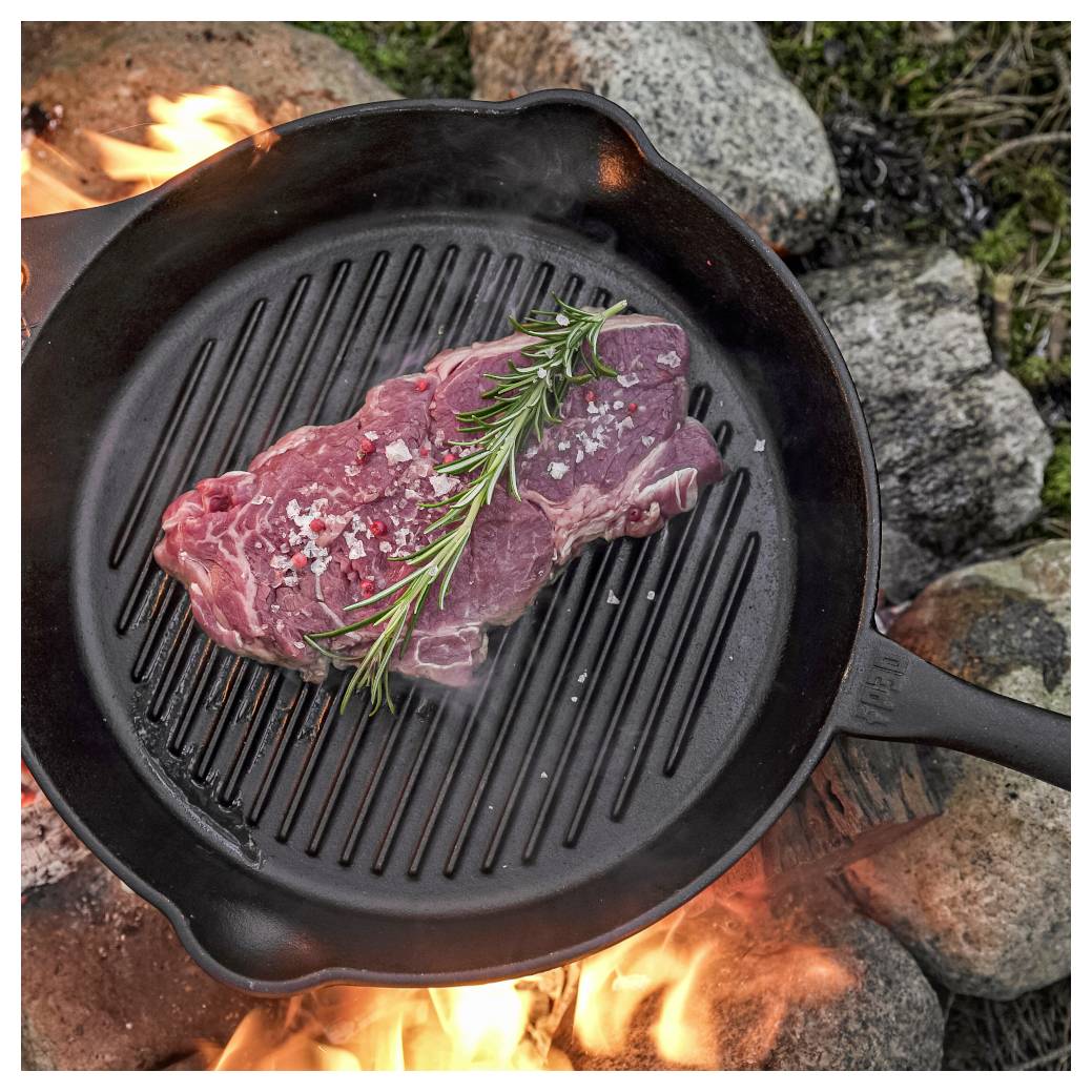 Rohes Steak mit Rosmarin, das in einer gusseisernen Pfanne über einem offenen Lagerfeuer zwischen Steinen angebraten wird.